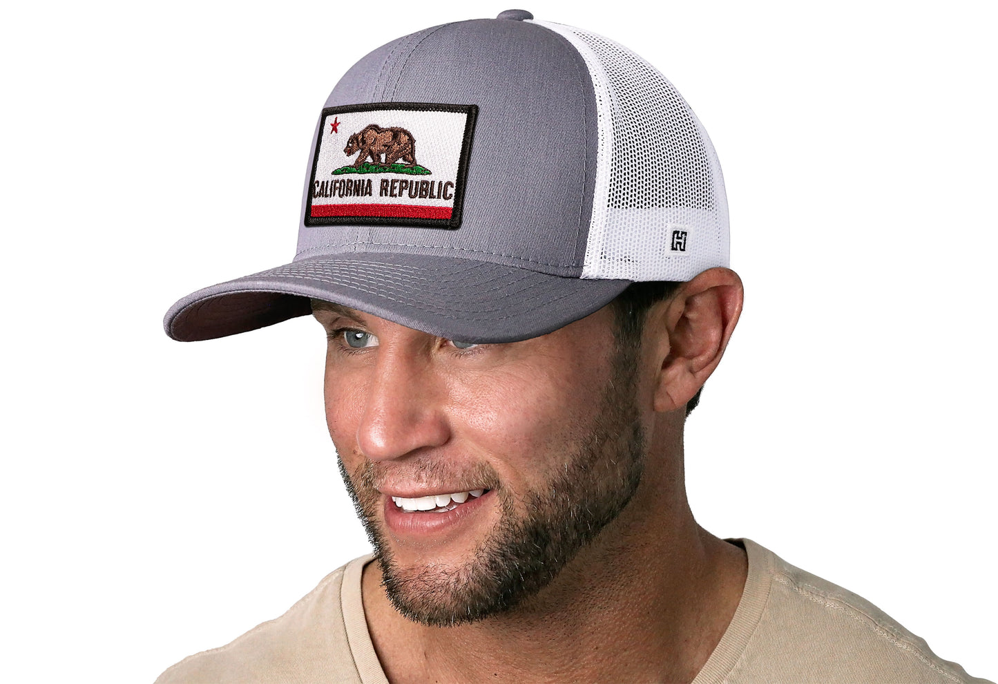 Gray-White trucker hat worn by a male model viewed from the left side and features a California Flag Embroidered Rectangle Patch displayed on the front panel. The hat has a classic mesh back design and a curved structured bill