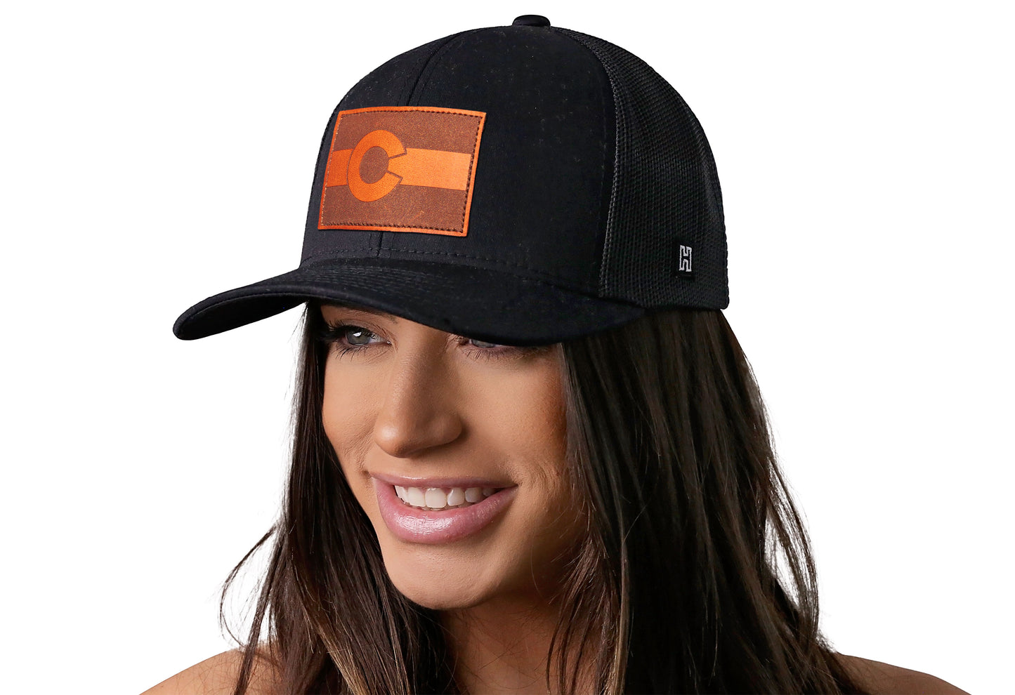 Black trucker hat worn by a female model viewed from the left side and features a Colorado Flag Leather Rectangle Patch displayed on the front panel. The hat has a classic mesh back design and a curved structured bill
