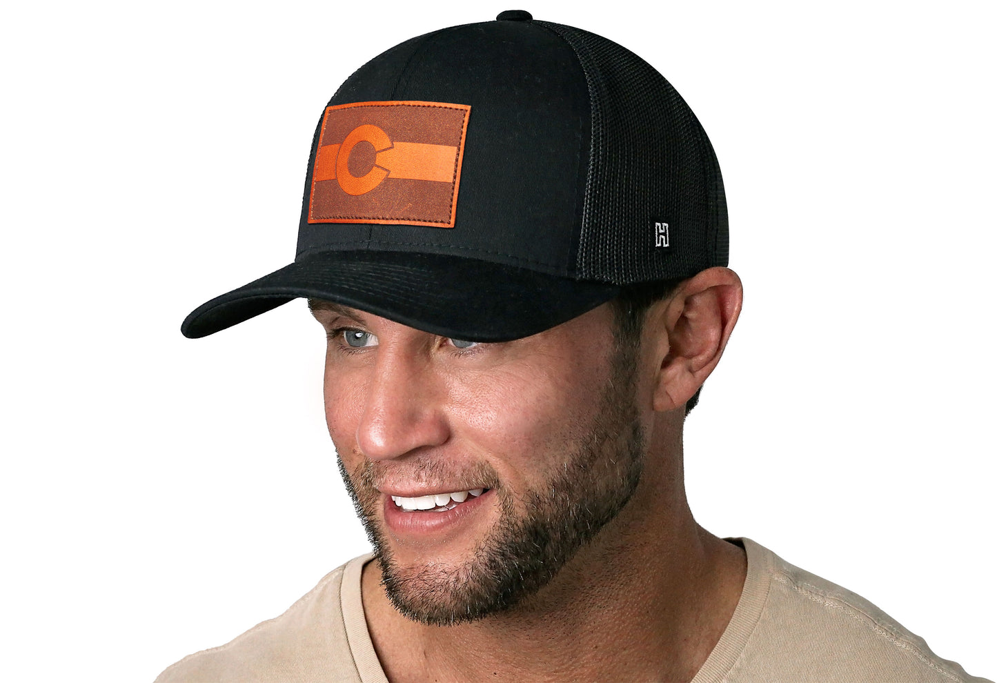 Black trucker hat worn by a male model viewed from the left side and features a Colorado Flag Leather Rectangle Patch displayed on the front panel. The hat has a classic mesh back design and a curved structured bill