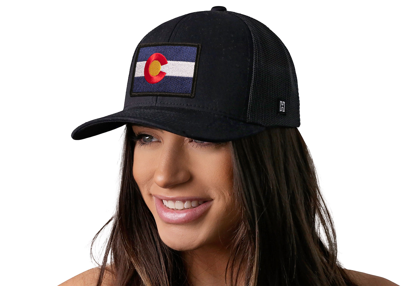 Black trucker hat worn by a female model viewed from the left side and features a Colorado Flag Embroidered Rectangle Patch displayed on the front panel. The hat has a classic mesh back design and a curved structured bill