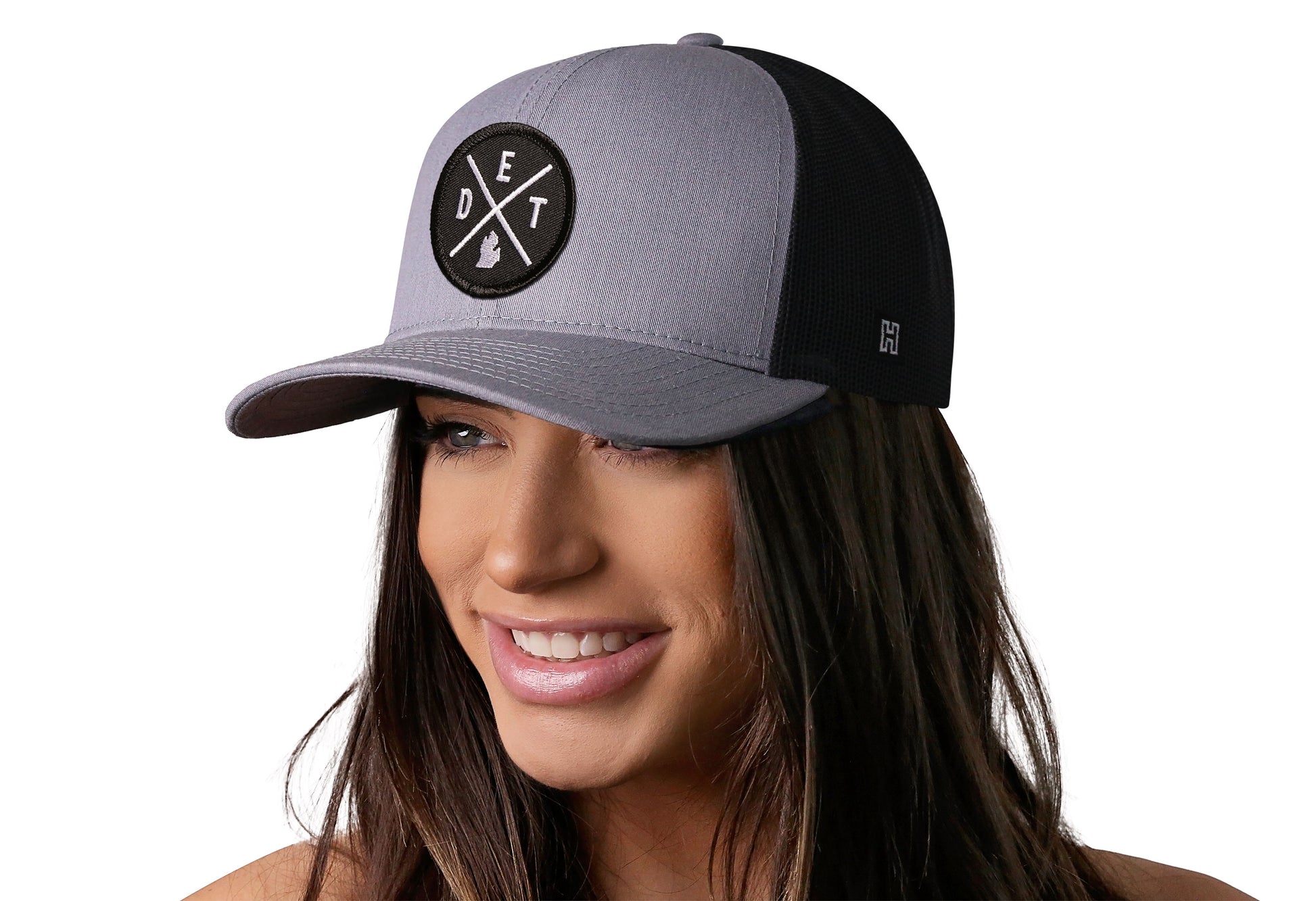 Gray-Black trucker hat worn by a female model viewed from the left side and features a Detroit DET X Embroidered Circle Patch displayed on the front panel. The hat has a classic mesh back design and a curved structured bill