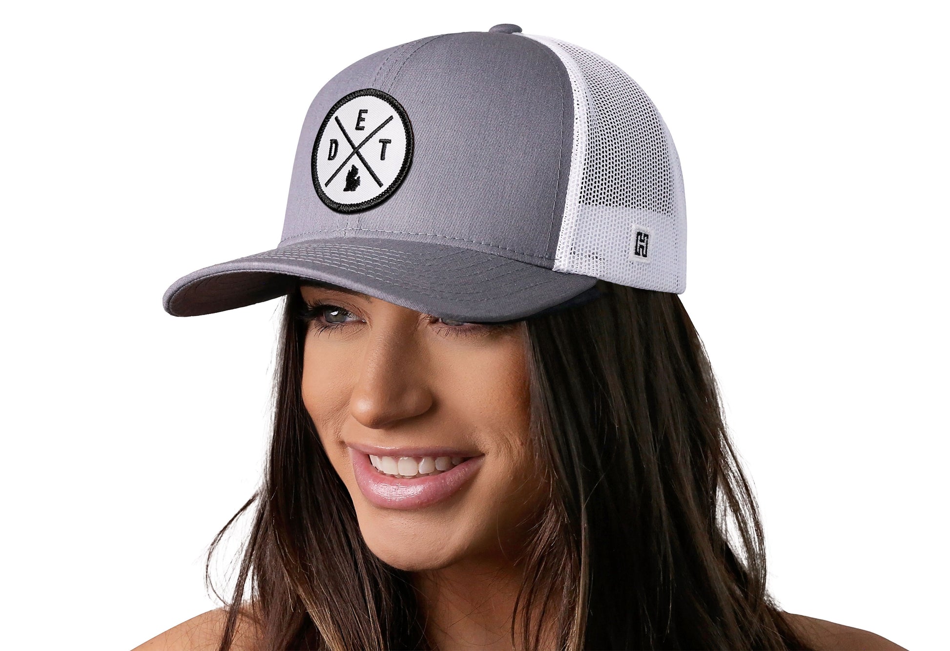 Gray-White trucker hat worn by a female model viewed from the left side and features a Detroit DET X Embroidered White Circle Patch displayed on the front panel. The hat has a classic mesh back design and a curved structured bill