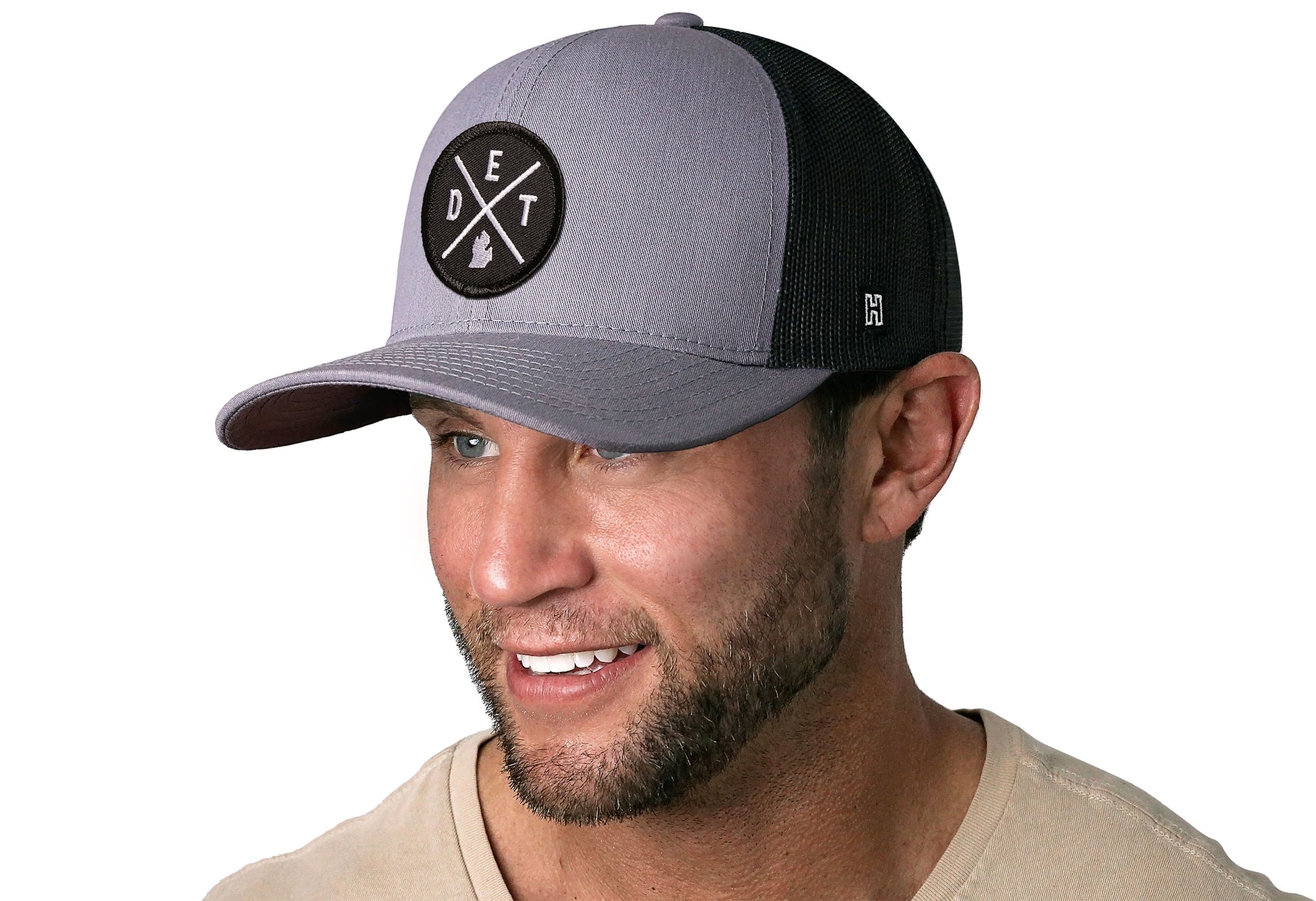 Gray-Black trucker hat worn by a male model viewed from the left side and features a Detroit DET X Embroidered Circle Patch displayed on the front panel. The hat has a classic mesh back design and a curved structured bill