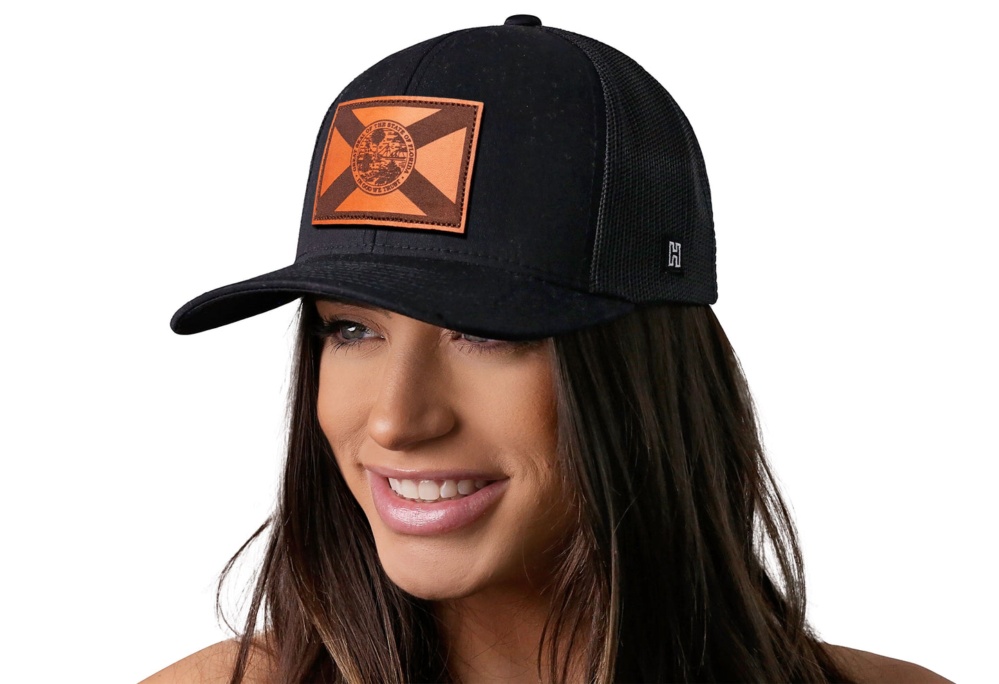 Black trucker hat worn by a female model viewed from the left side and features a Florida Flag Woven Leather Retangle Patch displayed on the front panel. The hat has a classic mesh back design and a curved structured bill