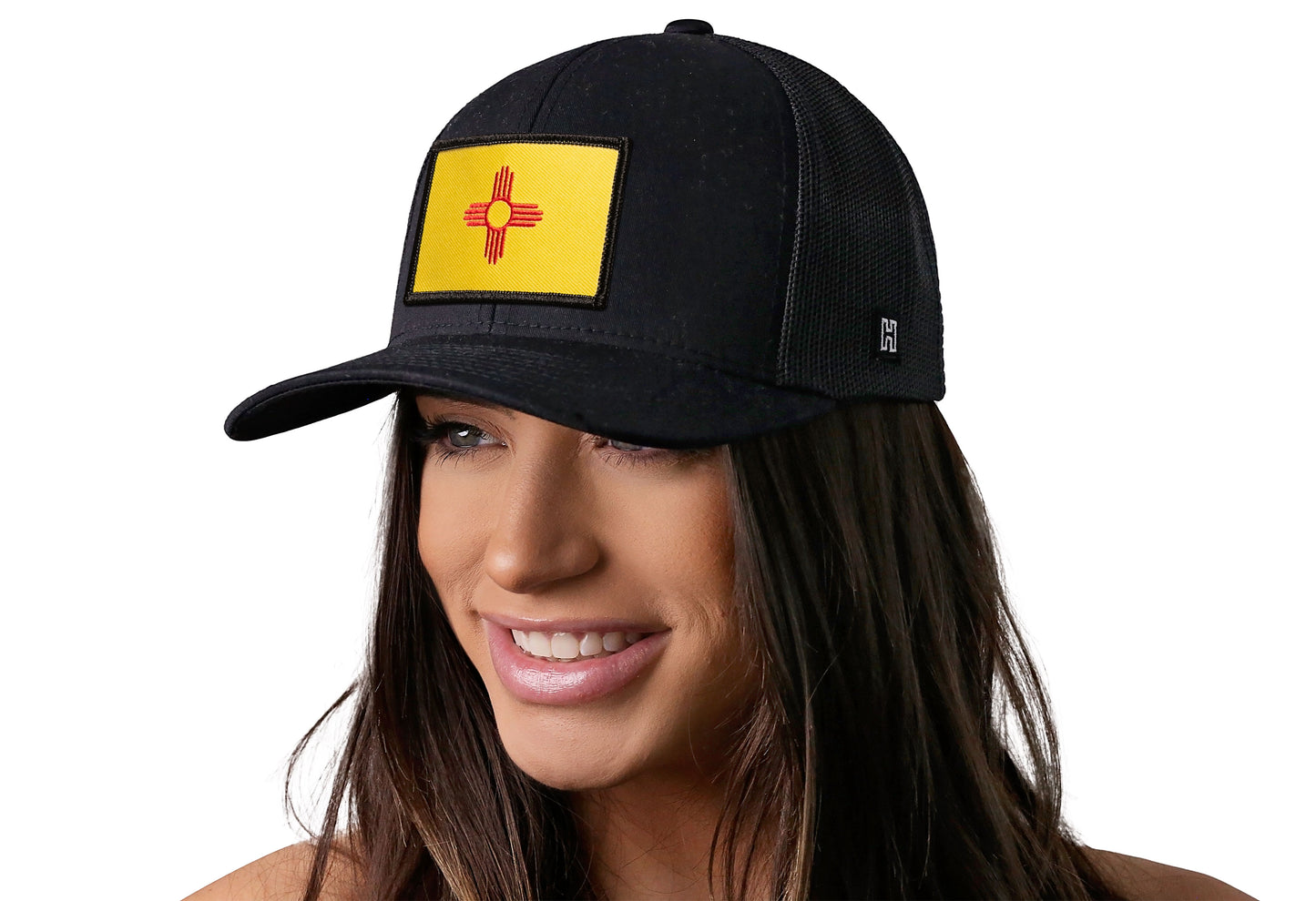 Black trucker hat worn by a female model viewed from the left side and features a New Mexico Woven Rectangle Patch displayed on the front panel. The hat has a classic mesh back design and a curved structured bill