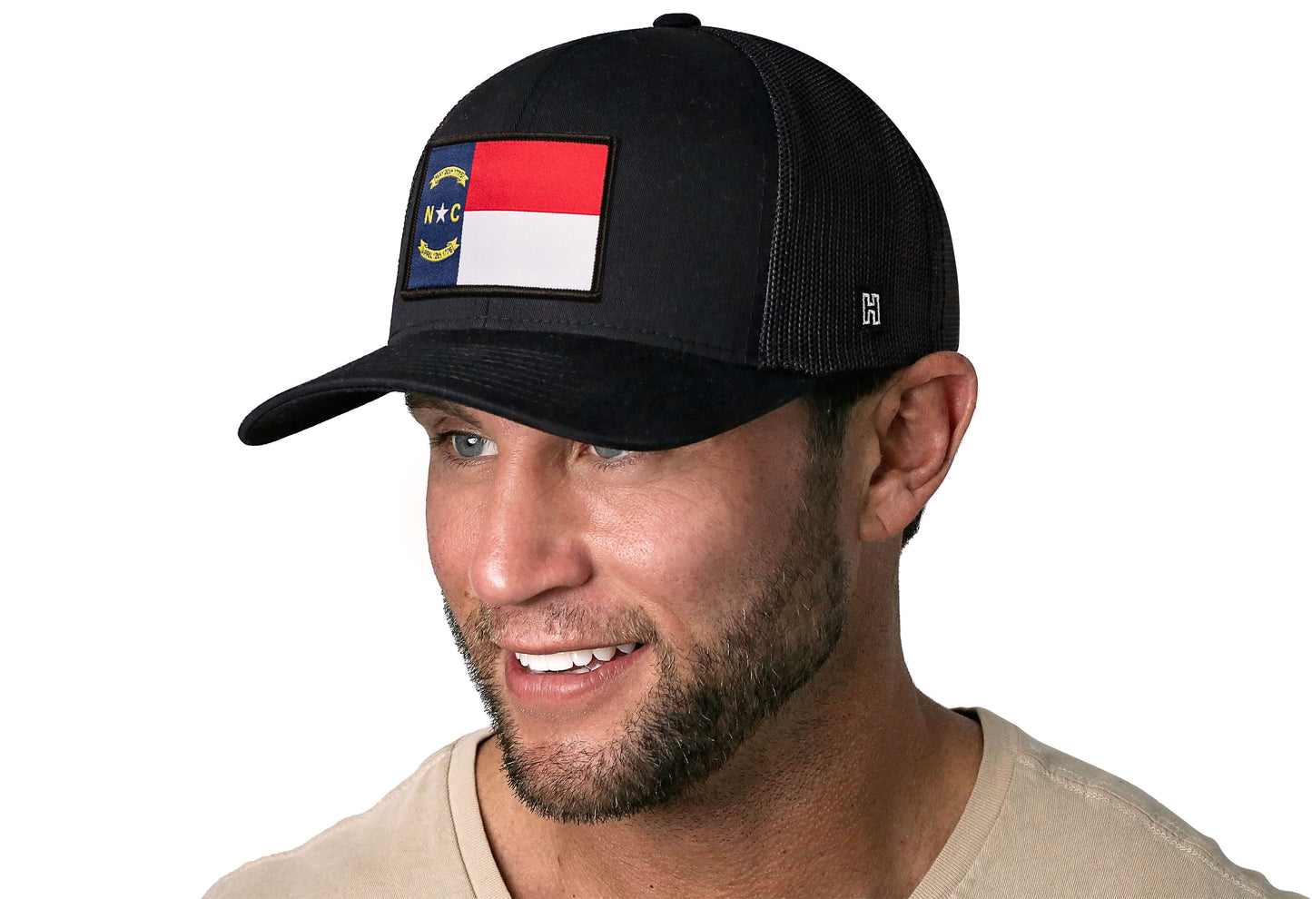 Black trucker hat worn by a male model viewed from the left side and features a North Carolina Flag Woven Rectangle Patch displayed on the front panel. The hat has a classic mesh back design and a curved structured bill