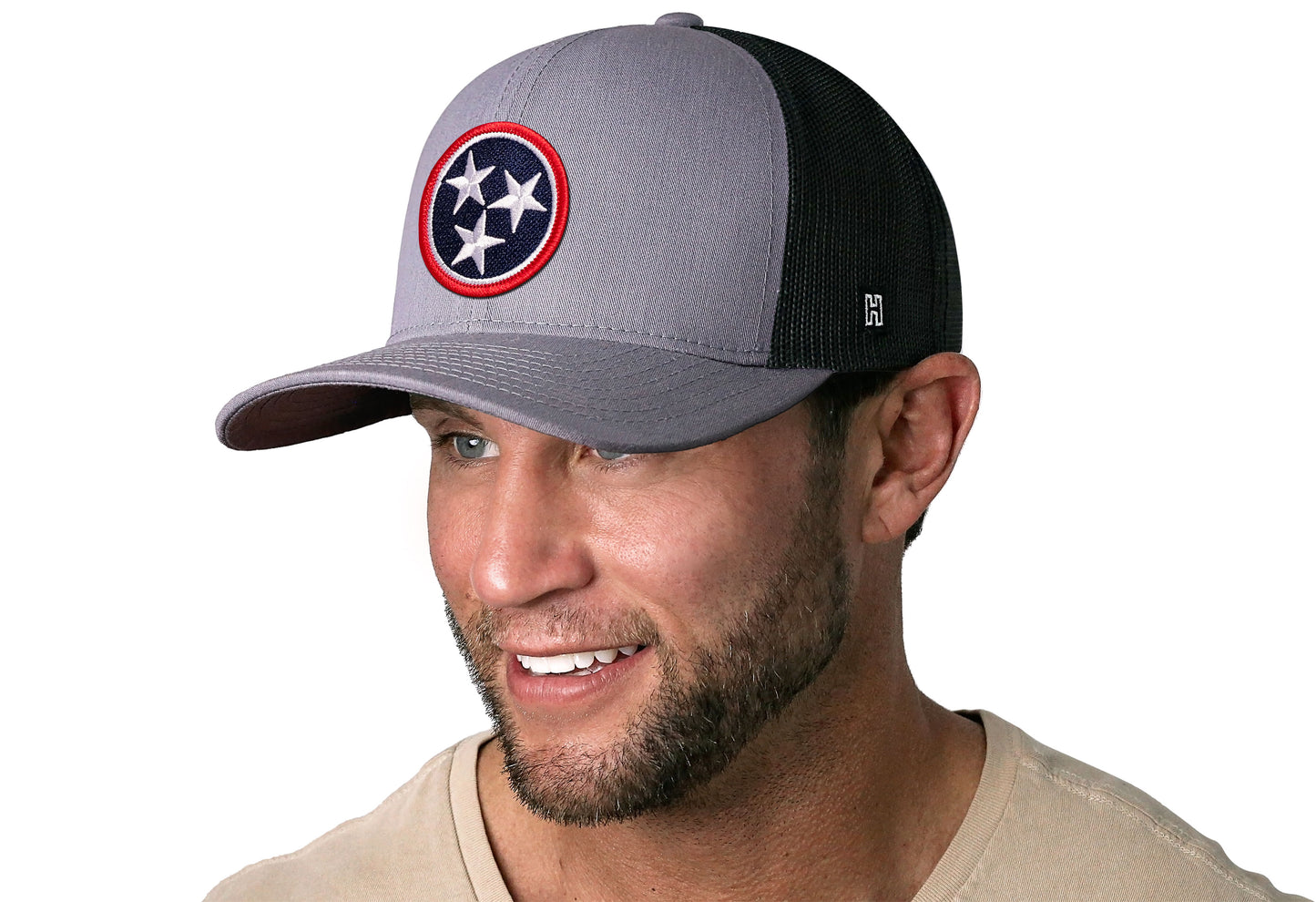 Gray-Black trucker hat worn by a male model viewed from the left side and features a Tennessee Gray- TN Embroidered Flag Patch displayed on the front panel. The hat has a classic mesh back design and a curved structured bill