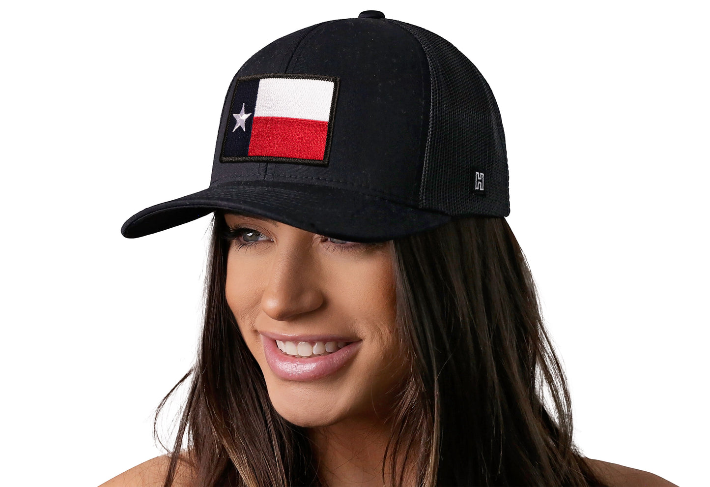 Black trucker hat worn by a female model viewed from the left side and features a Texas TX Embroidered Flag Patch displayed on the front panel. The hat has a classic mesh back design and a curved structured bill