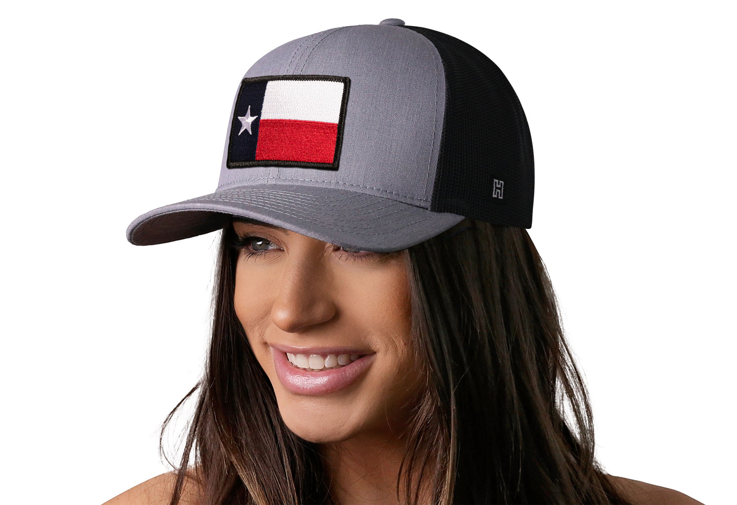 Gray-Black trucker hat worn by a female model viewed from the left side and features a Texas Gray-Black TX Embroidered Flag Patch displayed on the front panel. The hat has a classic mesh back design and a curved structured bill