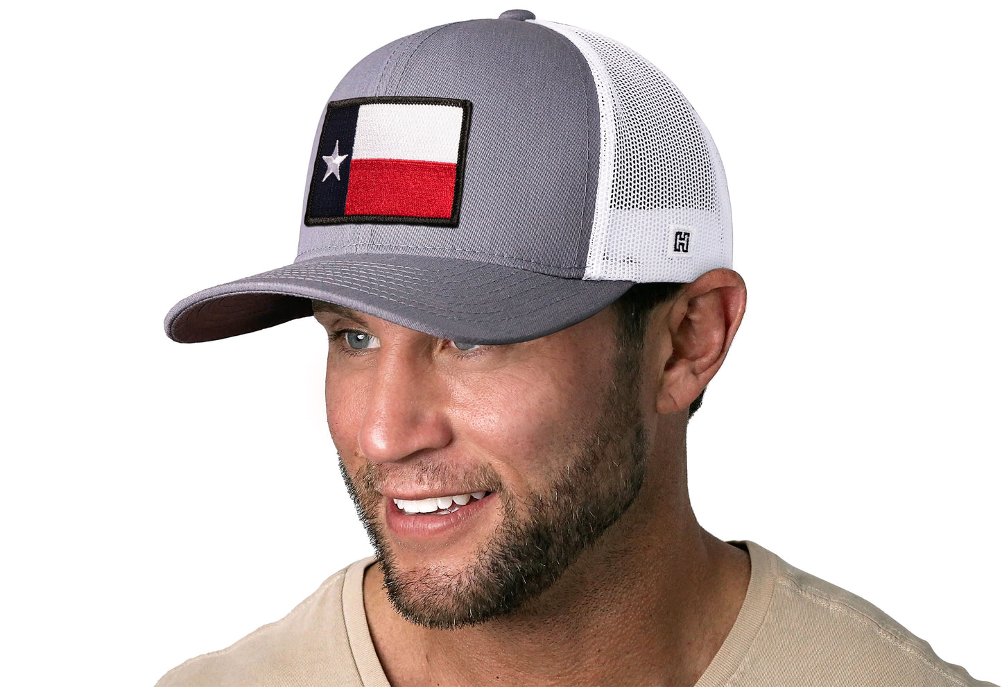 Gray-White trucker hat worn by a male model viewed from the left side and features a Texas TX Embroidered Flag Patch displayed on the front panel. The hat has a classic mesh back design and a curved structured bill