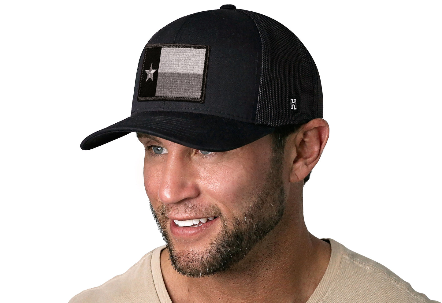 Black trucker hat worn by a male model viewed from the left side and features a Texas TX Tactical Embroidered Flag Patch displayed on the front panel. The hat has a classic mesh back design and a curved structured bill