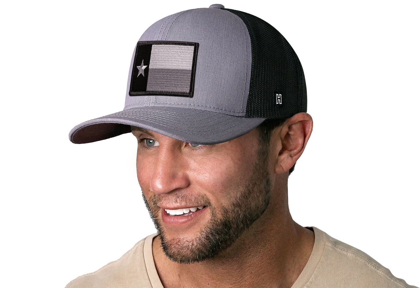 Gray-Black trucker hat worn by a male model viewed from the left side and features a Texas Gray-Black TX Tactical Embroidered Flag Patch displayed on the front panel. The hat has a classic mesh back design and a curved structured bill