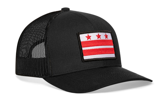 black trucker hat viewed from the right side, showcasing a bold red and white embroidered flag patch prominently displayed on the front