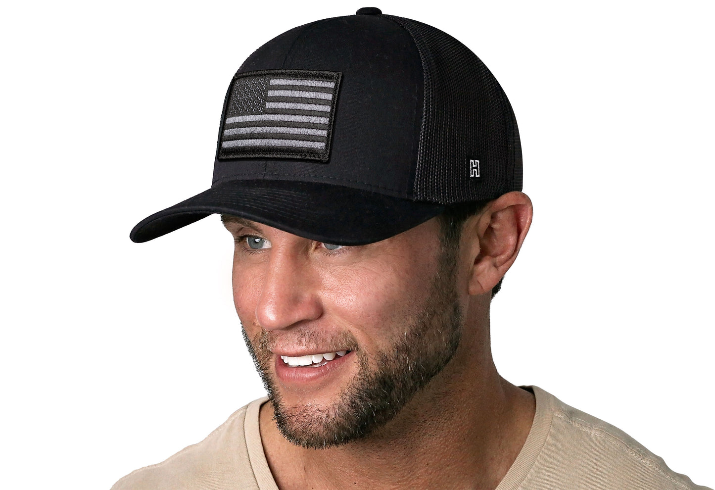 Black trucker hat worn by a male model viewed from the left side and features a American Flag Embroidered Rectangle Patch displayed on the front panel. The hat has a classic mesh back design and a curved structured bill