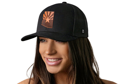 Black trucker hat worn by a female model viewed from the left side and features a Arizona Flag Leather Cut to Shape Flag Patch displayed on the front panel. The hat has a classic mesh back design and a curved structured bill
