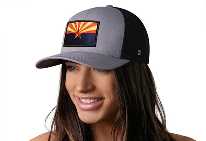 Gray-Black trucker hat worn by a female model viewed from the left side and features a Arizona Flag Embroidered Rectangle Patch displayed on the front panel. The hat has a classic mesh back design and a curved structured bill