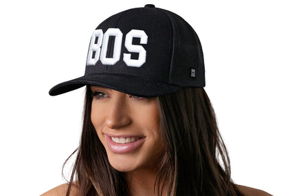 Black trucker hat worn by a female model viewed from the left side and features a BOS 3-D Puff Direct Embroidery displayed on the front panel. The hat has a classic mesh back design and a curved structured bill