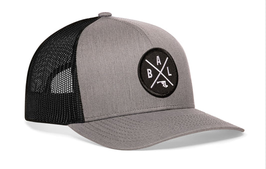 Gray-Black trucker hat viewed from the right side, showcasing a Baltimore BAL X Embroidered Circle Patch prominently displayed on the front