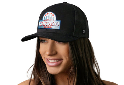 Black trucker hat worn by a female model viewed from the left side and features a Chicago Cityscape Embroidered Patch displayed on the front panel. The hat has a classic mesh back design and a curved structured bill