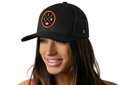 black trucker hat worn by a female model viewed from the left side and features a bold orange and black embroidered X patch displayed on the front panel. The hat has a classic mesh back design and a curved structured bill