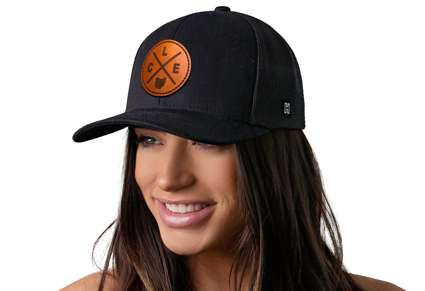 Black trucker hat worn by a female model viewed from the left side and features a Cleveland CLE X Leather Circle Patch displayed on the front panel. The hat has a classic mesh back design and a curved structured bill