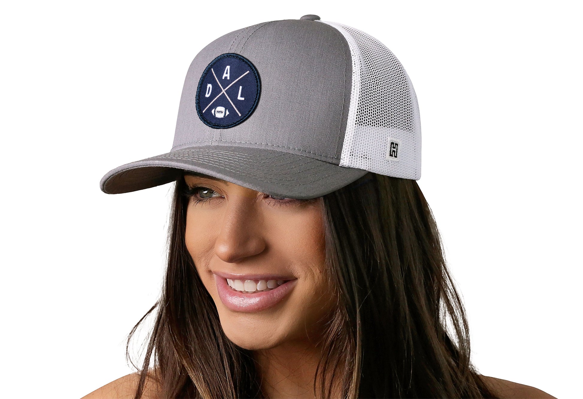 gray-white trucker hat worn by a female model viewed from the left side and features a bold navy, silver, and white embroidered X patch displayed on the front panel. The hat has a classic mesh back design and a curved structured bill