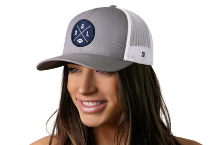 gray-white trucker hat worn by a female model viewed from the left side and features a bold navy, silver, and white embroidered X patch displayed on the front panel. The hat has a classic mesh back design and a curved structured bill