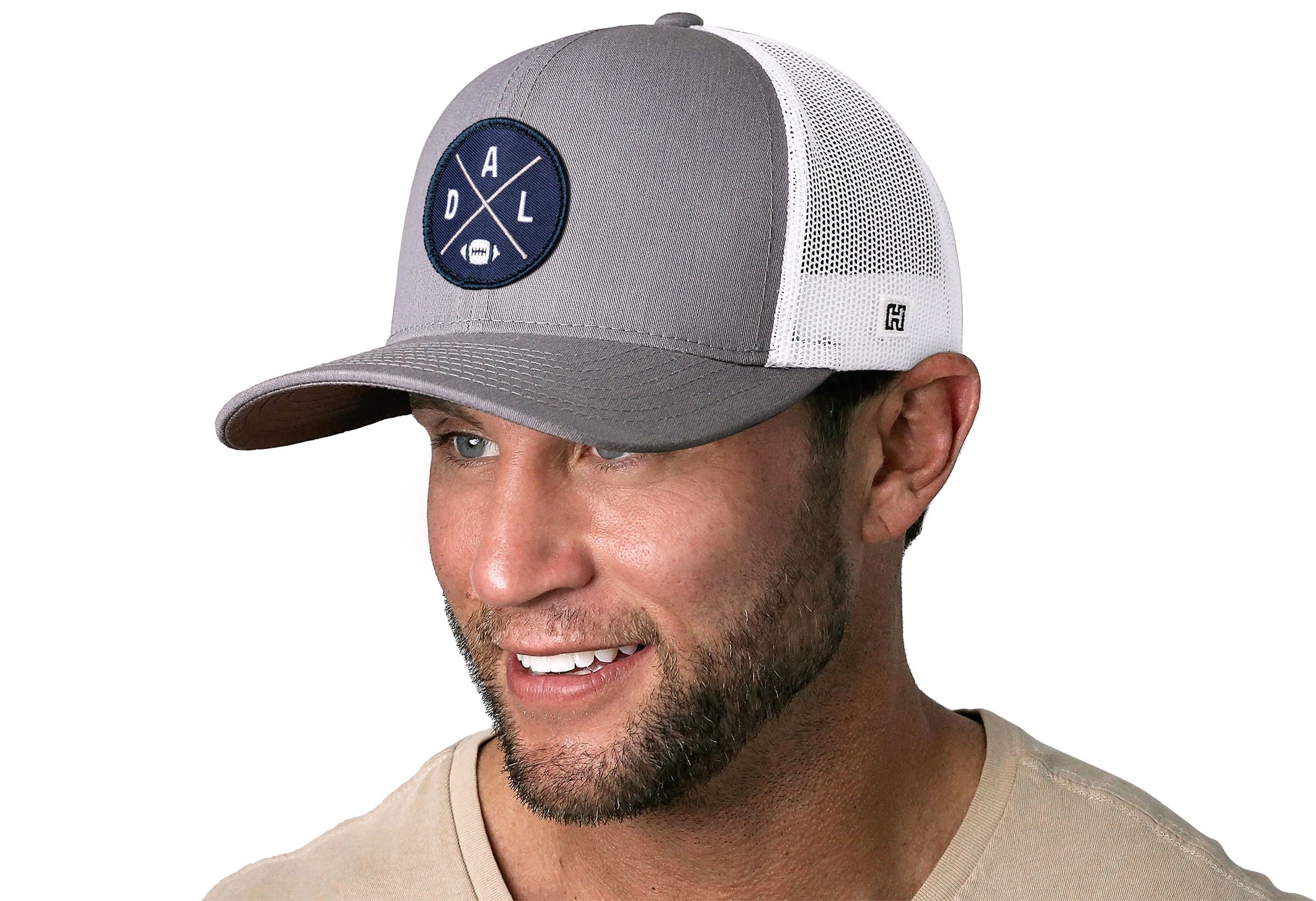 gray-white trucker hat worn by a male model viewed from the left side and features a bold navy, silver, and white embroidered X patch displayed on the front panel. The hat has a classic mesh back design and a curved structured bill