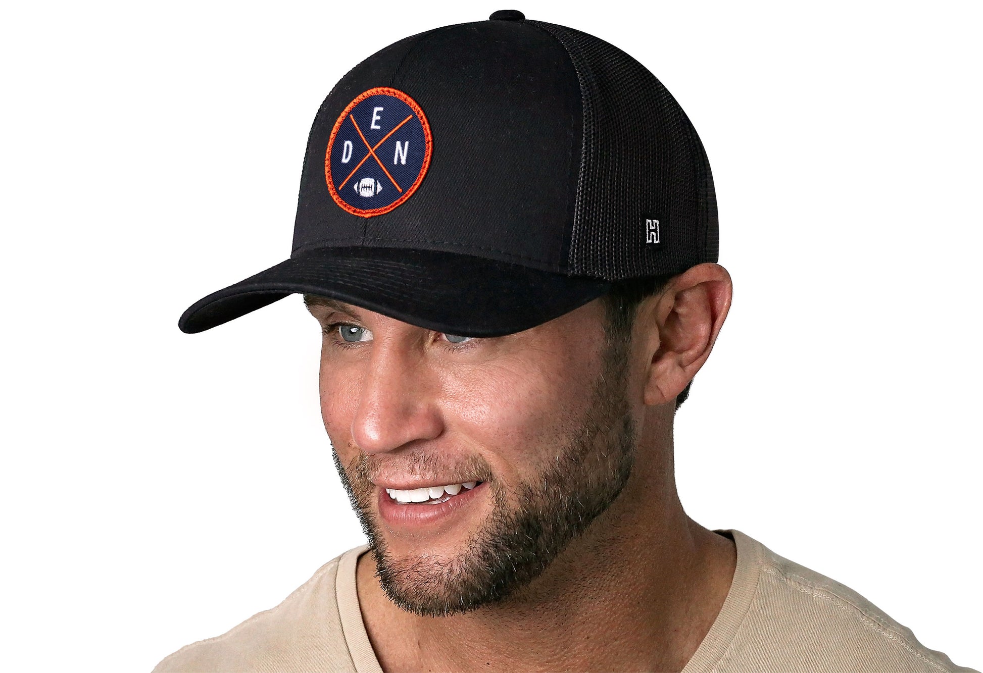 black trucker hat worn by a male model viewed from the left side and features a bold orange and navy embroidered X patch displayed on the front panel. The hat has a classic mesh back design and a curved structured bill