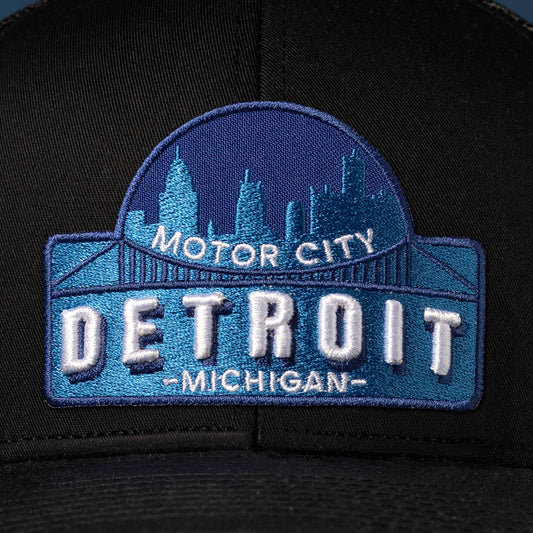 Black trucker hat viewed from the right side, showcasing a Detroit Cityscape Embroidered Patch prominently displayed on the front