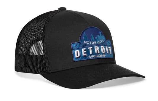 Black trucker hat worn by a male model viewed from the left side and features a Detroit Cityscape Embroidered Patch displayed on the front panel. The hat has a classic mesh back design and a curved structured bill