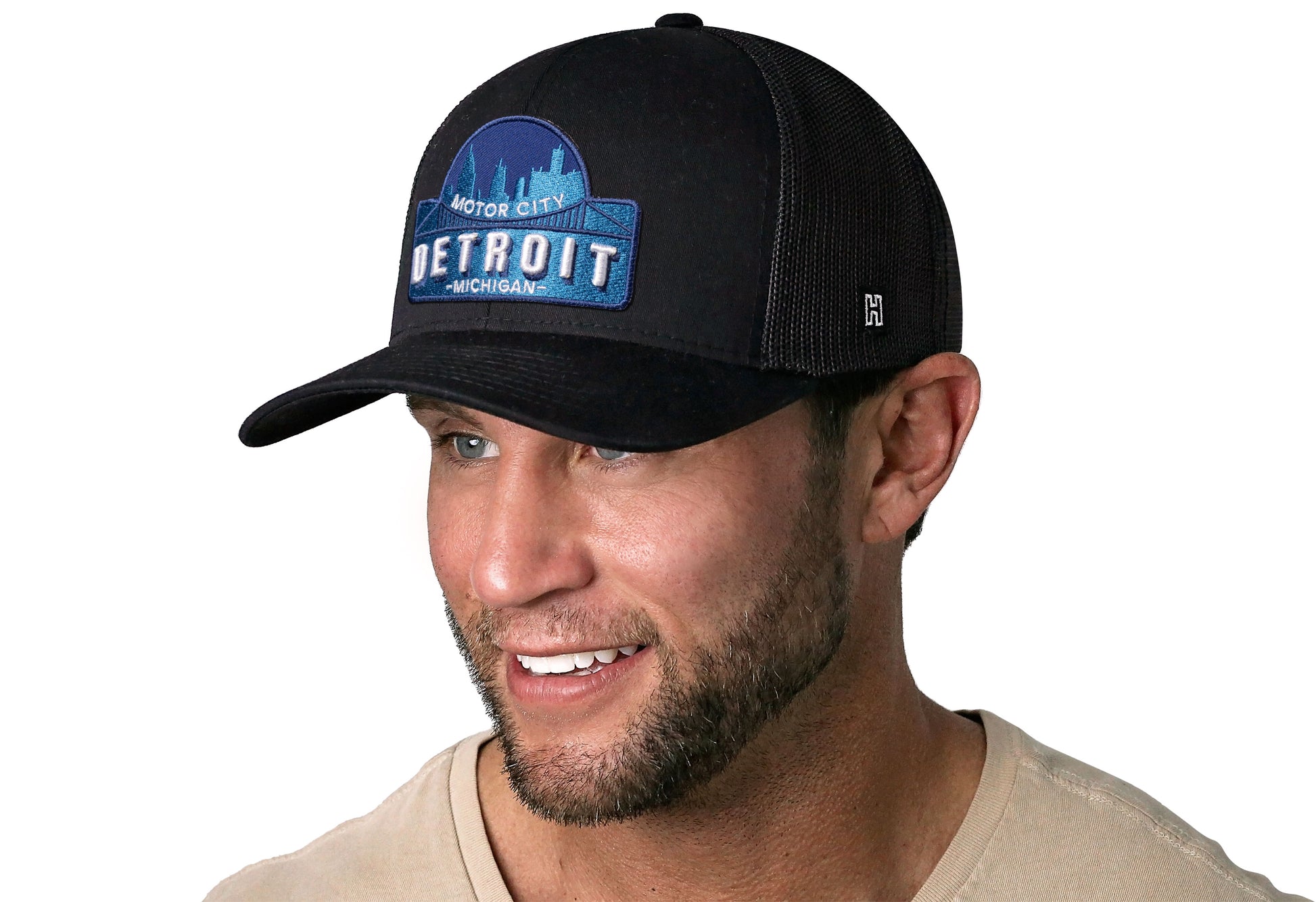 Black trucker hat worn by a male model viewed from the left side and features a Detroit Cityscape Embroidered Patch displayed on the front panel. The hat has a classic mesh back design and a curved structured bill