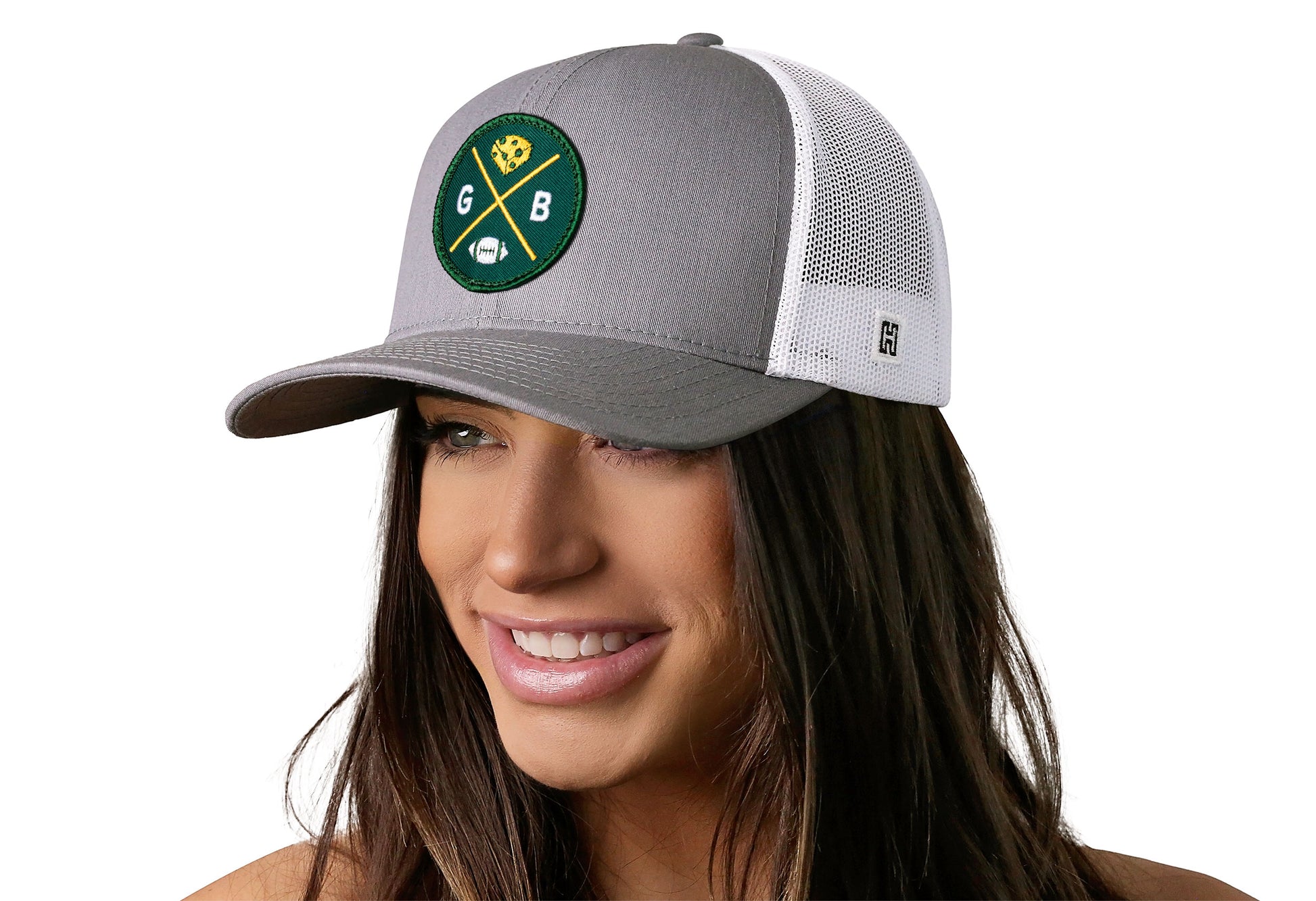 gray-white trucker hat worn by a female model viewed from the left side and features a bold dark green and gold embroidered X patch displayed on the front panel. The hat has a classic mesh back design and a curved structured bill