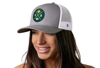 gray-white trucker hat worn by a female model viewed from the left side and features a bold dark green and gold embroidered X patch displayed on the front panel. The hat has a classic mesh back design and a curved structured bill