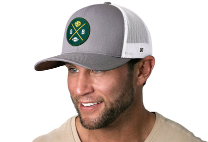 gray-white trucker hat worn by a male model viewed from the left side and features a bold dark green and gold embroidered X patch displayed on the front panel. The hat has a classic mesh back design and a curved structured bill