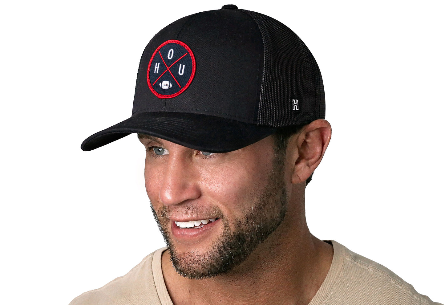 black trucker hat worn by a male model viewed from the left side and features a bold blue and red embroidered X patch displayed on the front panel. The hat has a classic mesh back design and a curved structured bill