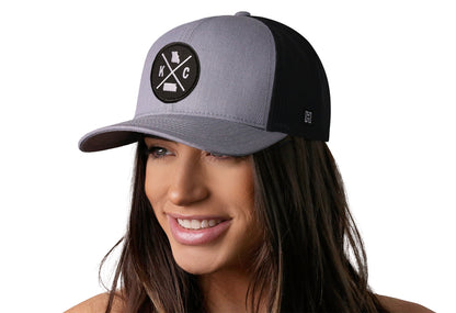 Gray-Black trucker hat worn by a female model viewed from the left side and features a Kansas City KC X Embroidered Circle Patch displayed on the front panel. The hat has a classic mesh back design and a curved structured bill