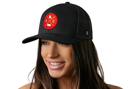 Black trucker hat worn by a female model viewed from the left side and features a Kansas City KC X Embroidered Red, White, Gold Circle Patch displayed on the front panel. The hat has a classic mesh back design and a curved structured bill