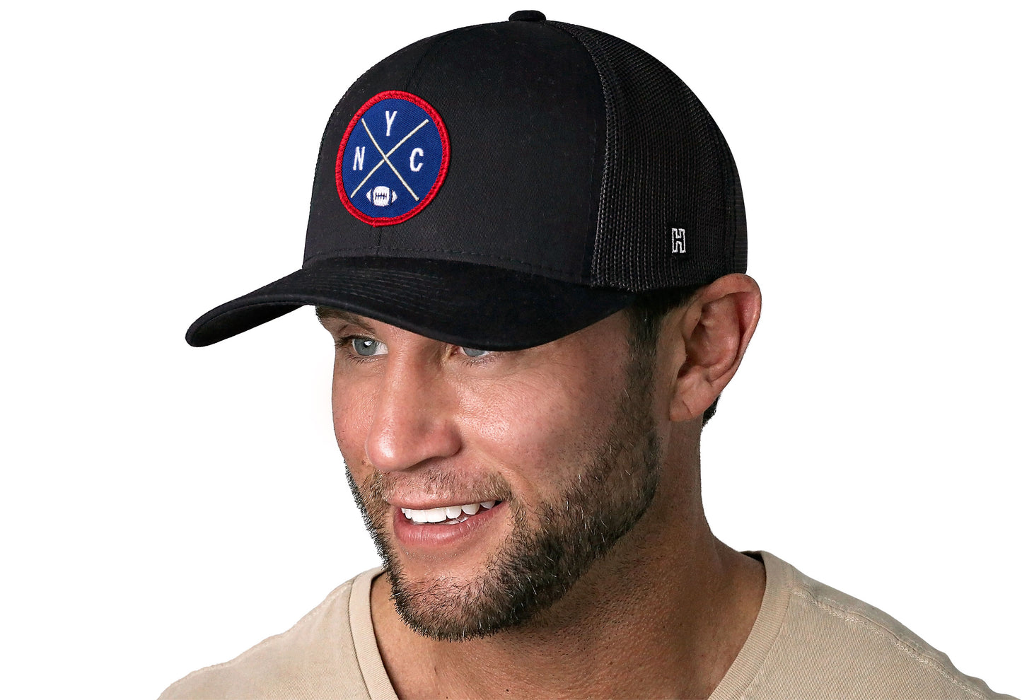 black trucker hat worn by a male model viewed from the left side and features a bold blue and red embroidered X patch displayed on the front panel. The hat has a classic mesh back design and a curved structured bill