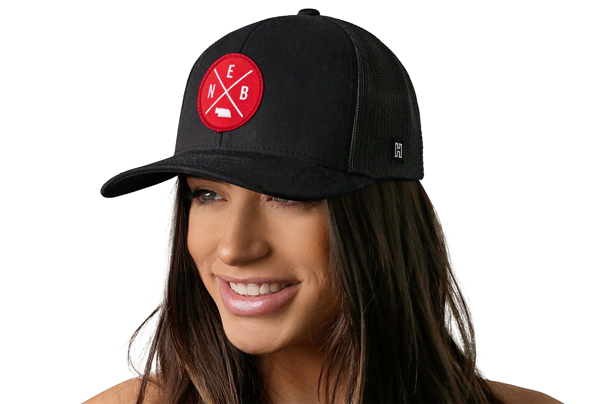 black trucker hat worn by a female model viewed from the left side and features a bold red and white embroidered X patch displayed on the front panel. The hat has a classic mesh back design and a curved structured bill