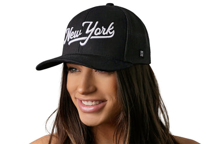 black trucker hat worn by a female model viewed from the left side and features a White 'New York' Puff Script 3D Embroidery displayed on the front panel. The hat has a classic mesh back design and a curved structured bill