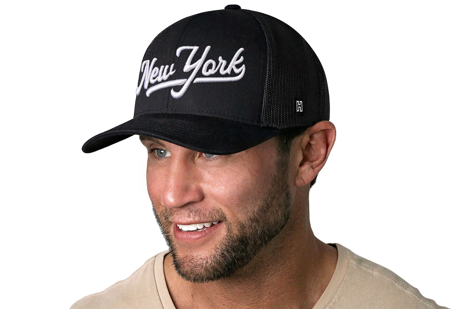 black trucker hat worn by a male model viewed from the left side and features a White 'New York' Puff Script 3D Embroidery displayed on the front panel. The hat has a classic mesh back design and a curved structured bill