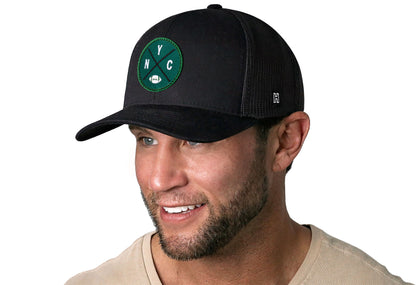 black trucker hat worn by a male model viewed from the left side and features a bold green and white embroidered X patch displayed on the front panel. The hat has a classic mesh back design and a curved structured bill