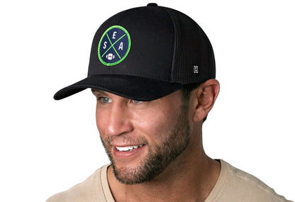 black trucker hat worn by a male model viewed from the left side and features a bold navy and lime green embroidered X patch displayed on the front panel. The hat has a classic mesh back design and a curved structured bill