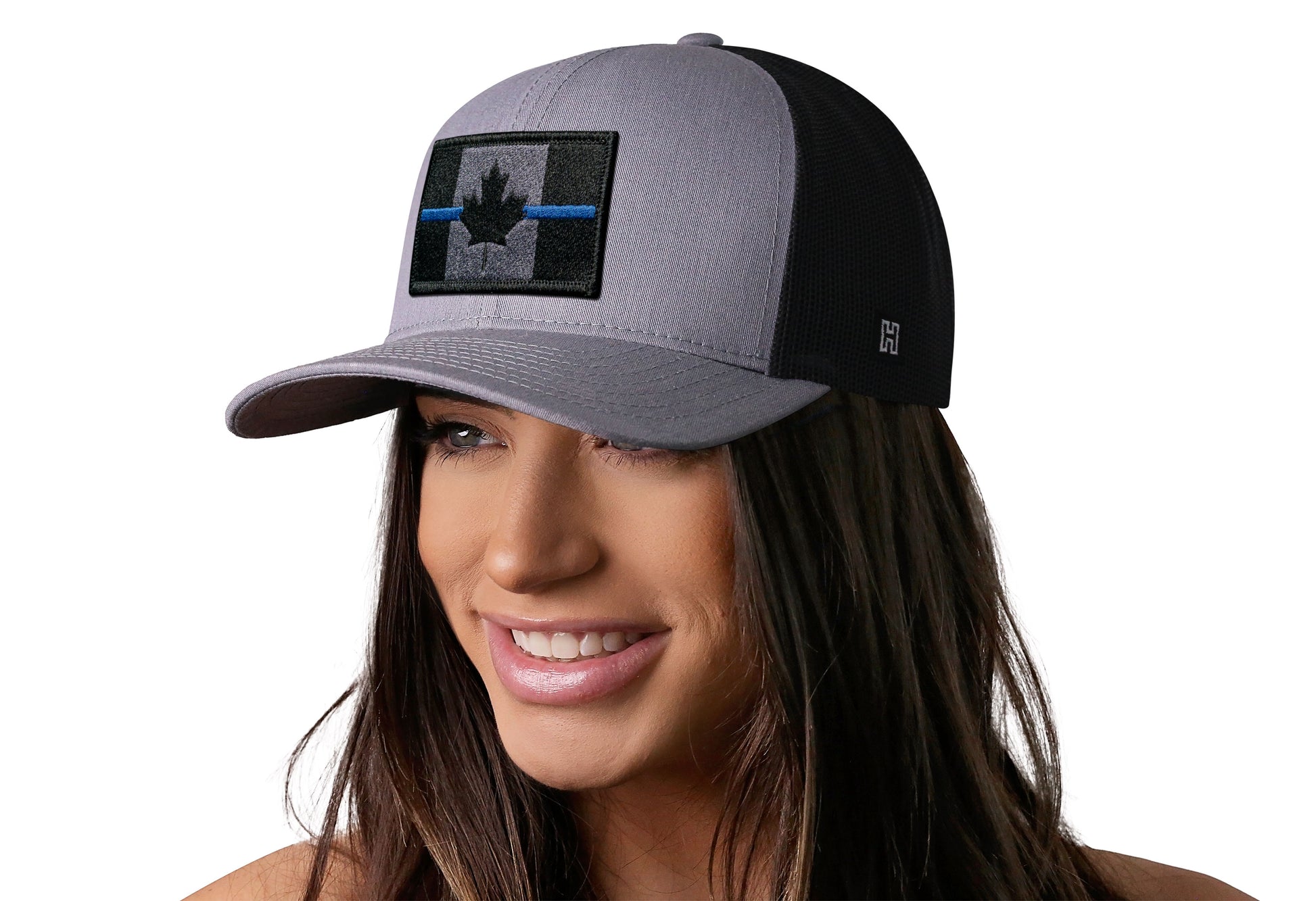 Gray-Black trucker hat worn by a female model viewed from the left side and features a Thin Blue Canadian Flag Embroidered Rectangle Patch displayed on the front panel. The hat has a classic mesh back design and a curved structured bill