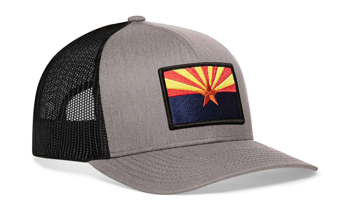 gray-black trucker hat viewed from the right side, showcasing a bold red, yellow and blue embroidered flag patch prominently displayed on the front