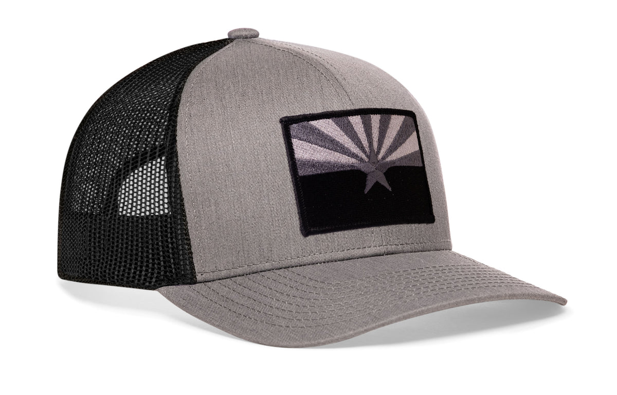 gray-black trucker hat viewed from the right side, showcasing a bold black and gray embroidered flag patch prominently displayed on the front