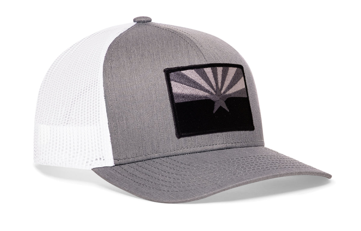 gray-white trucker hat viewed from the right side, showcasing a bold black and gray embroidered flag patch prominently displayed on the front