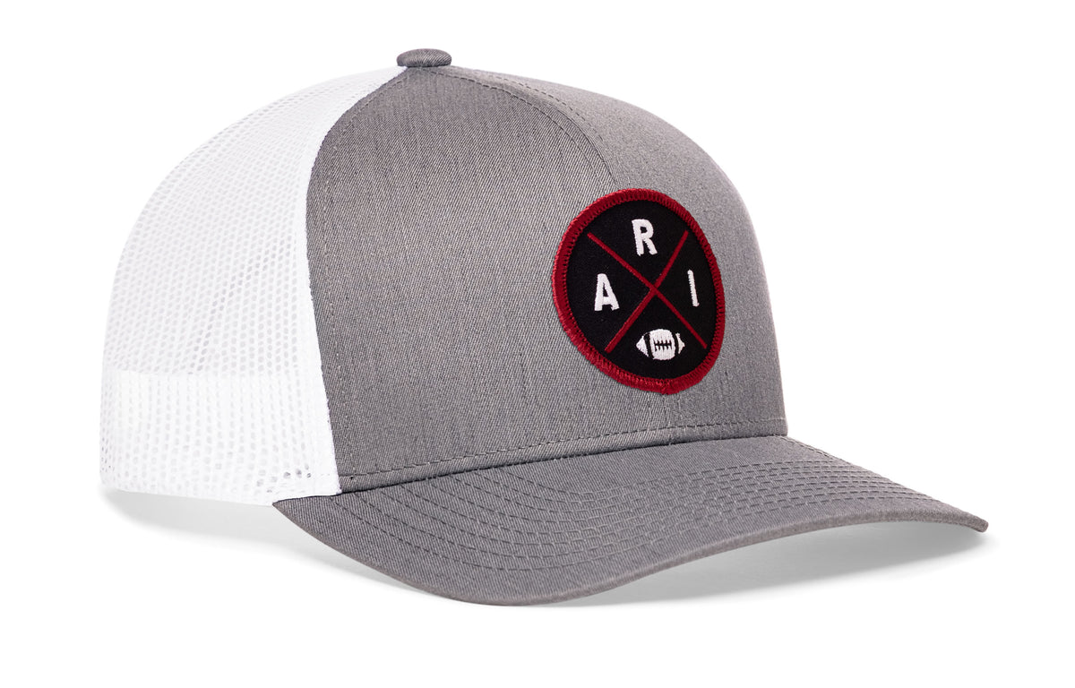 gray-white trucker hat viewed from the right side, showcasing a bold red and black embroidered X patch prominently displayed on the front