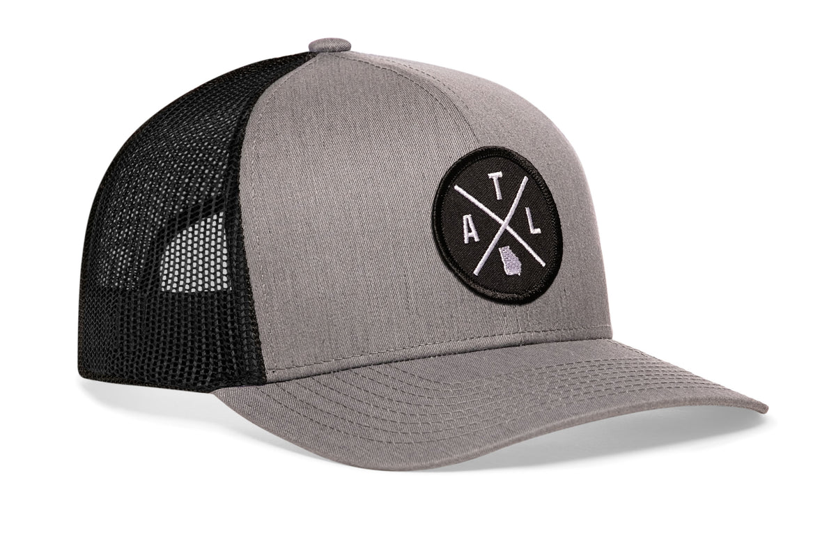 gray-black trucker hat viewed from the right side, showcasing a bold black and white embroidered X patch prominently displayed on the front